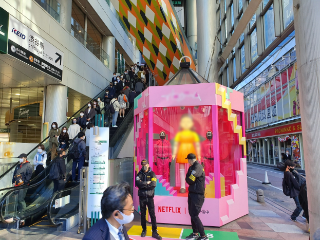 Eine Gruppe von Menschen steht vor einem Gebäude mit Glaswänden, Säulen und Texttafeln, einige tragen Masken, während hinter ihnen eine Rolltreppe mit Menschen nach oben führt; ein pinker Kasten in der Mitte zeigt Schaufensterpuppen und trägt den Text "Netflix's new Netflix booth in Tokyo."