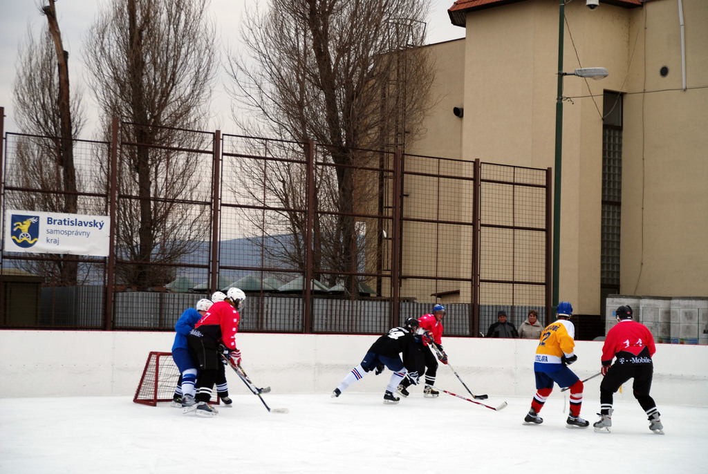 Menschen beim Eishockeyspielen auf einer Eisfläche mit Gebäuden, Bäumen, einer Straßenlaterne, einem Namensschild und Zäunen im Hintergrund unter einem klaren Himmel.