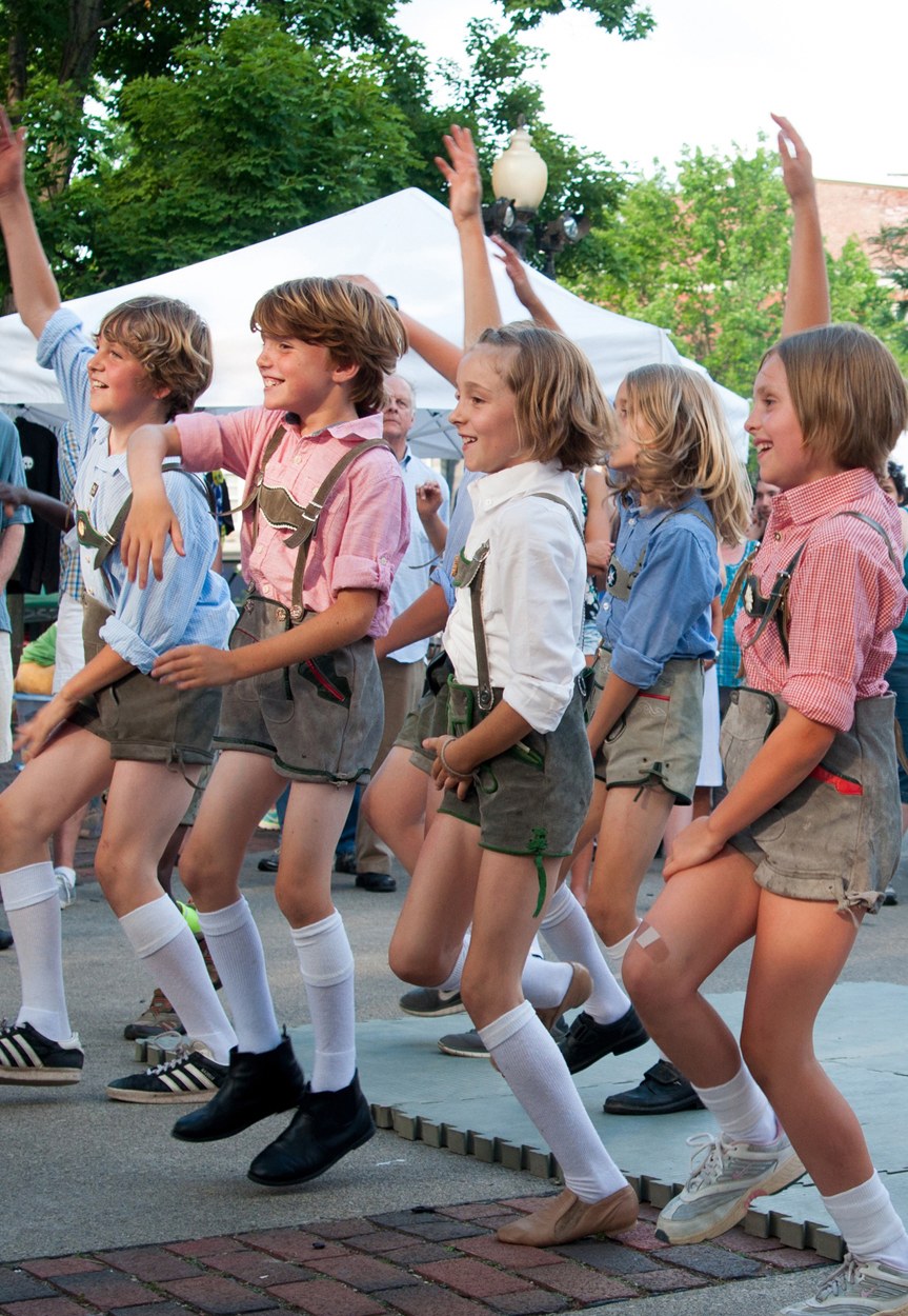 Kinder in traditioneller bayrischer Tracht, die auf einer Straße während eines Outdoor-Festivals tanzen, umgeben von Zuschauern, Zelten, Gebäuden und einem klaren blauen Himmel.