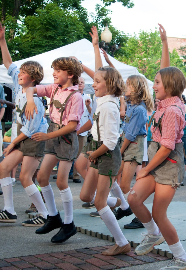 Kinder in traditioneller bayrischer Tracht, die auf einer Straße während eines Outdoor-Festivals tanzen, umgeben von Zuschauern, Zelten, Gebäuden und einem klaren blauen Himmel.