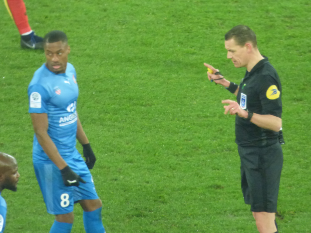 Ein Schiedsrichter spricht mit einem Fußballspieler auf dem Feld, umgeben von einer Gruppe von Menschen, wobei eine Person ein Objekt hält und die Beine einer anderen Person im Hintergrund zu sehen sind.