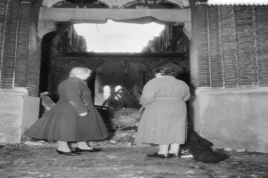 Zwei Frauen stehen in der Tür eines feuerbeschädigten Gebäudes, umgeben von verkohlten Überresten, auf einem Schwarz-Weiß-Bild.