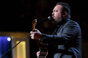 Ein Mann in einem Anzug spielt Gitarre und singt in ein Mikrofon auf der Bühne, mit Dierks Bentley im Hintergrund.