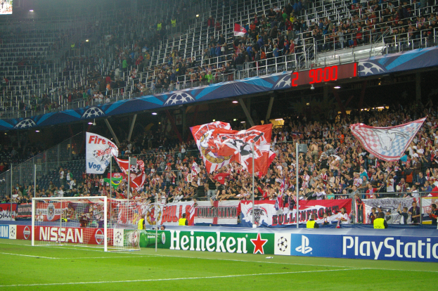 Ein Fußballspiel wird in einem gut beleuchteten Stadion mit einer großen Zuschauermenge, grünem Rasen, einem mit Bannern geschmückten Tor und einem Bildschirm in der oberen linken Ecke gespielt.