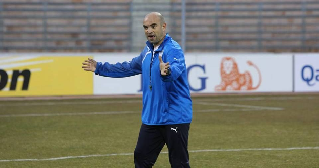Ein Mann in einer blauen Jacke steht auf einem Fußballfeld während eines Trainings, schaut nach vorne und hat einen entschlossenen Gesichtsausdruck, mit Tafeln im Hintergrund, die Text und Logos anzeigen.
