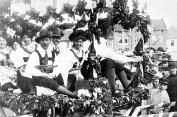 Eine Gruppe von Menschen in traditioneller bayrischer Tracht, einige mit Musikinstrumenten, fährt auf einemblattgeschmückten Festwagen in einem Umzug, mit Gebäuden und einem klaren Himmel im Hintergrund, dargestellt in Schwarz-Weiß.