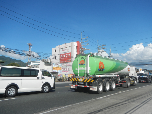 Ein großer Tanklastzug fährt auf einer Straße neben einem weißen Transporter, mit Strommasten, Gebäuden, Plakaten, Bäumen, Hügeln und einem bewölkten Himmel im Hintergrund.
