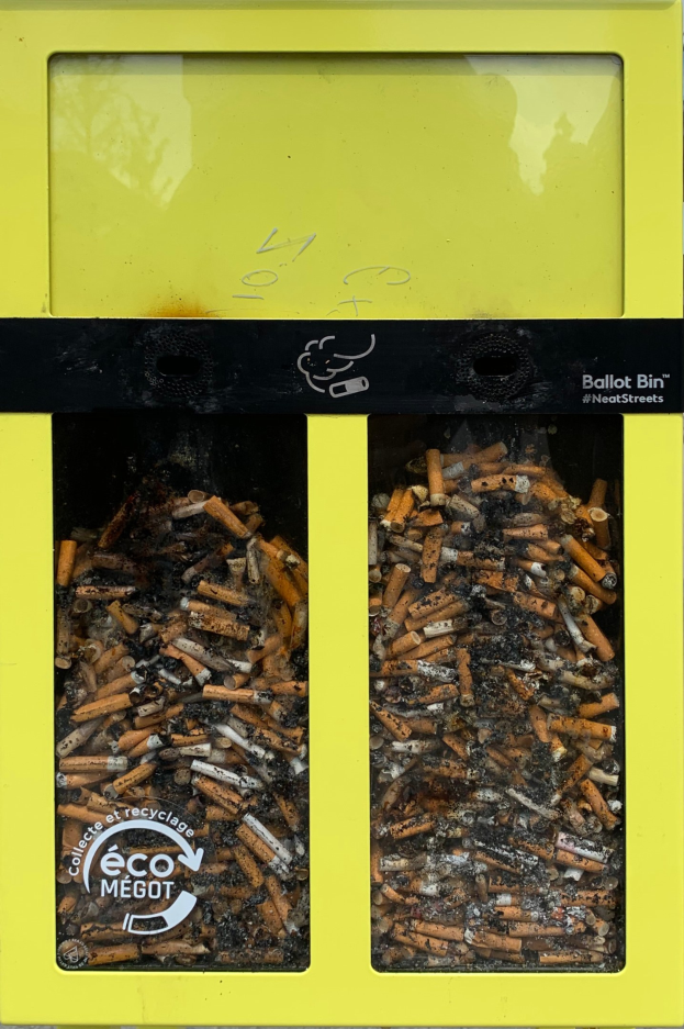 Gelber Müllcontainer voller Zigarettenstummel auf einem Gehweg, mit Text auf dem Container.