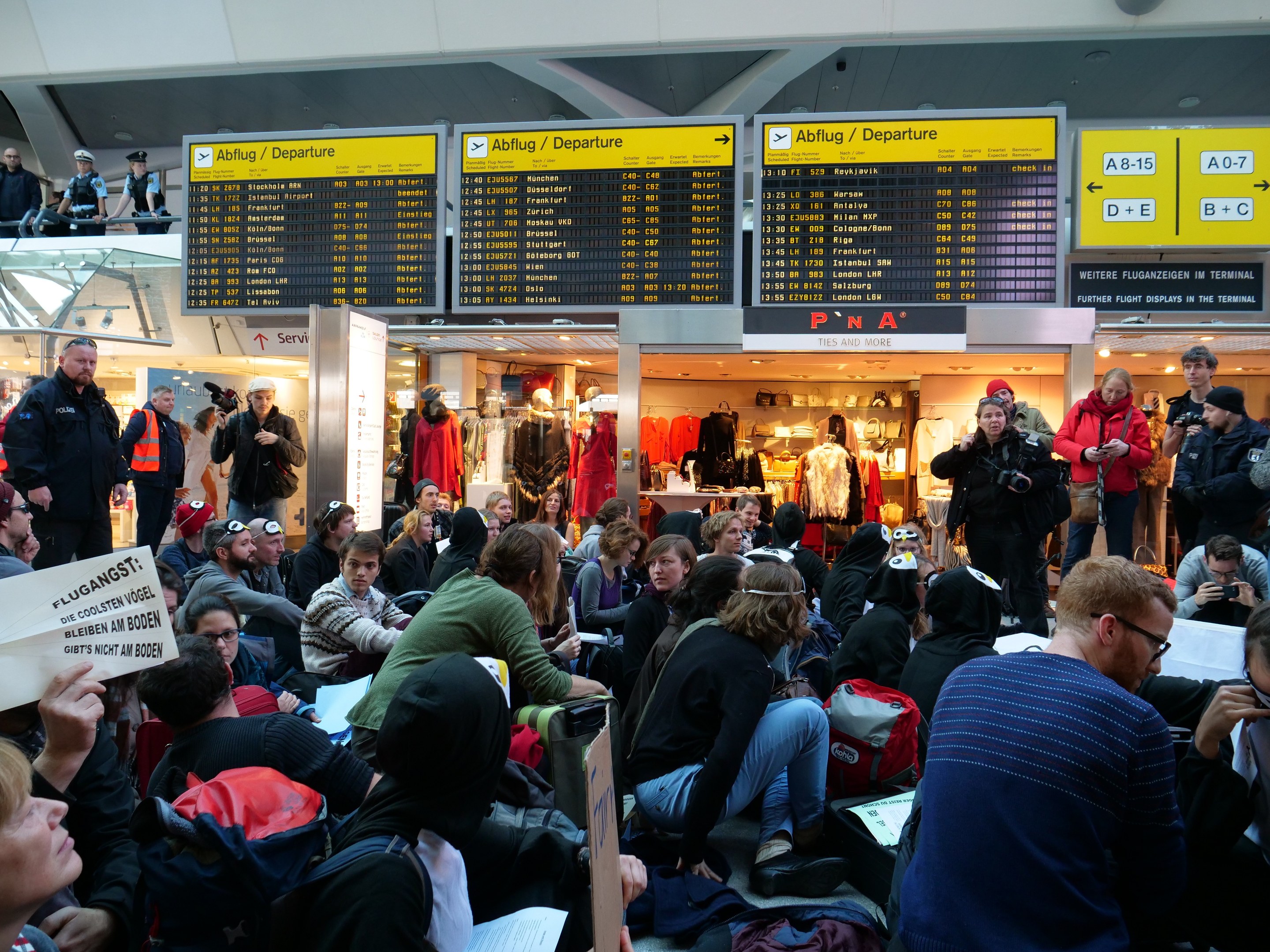 Eine große Gruppe von Menschen an einem Flughafen, einige sitzen mit Taschen und Papieren, andere stehen, mit Texttafeln, Schaufensterpuppen in Kleidern und Deckenbeleuchtung im Hintergrund, was auf eine Protestaktion hindeutet.