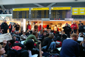 Eine große Gruppe von Menschen an einem Flughafen, einige sitzen mit Taschen und Papieren, andere stehen, mit Texttafeln, Schaufensterpuppen in Kleidern und Deckenbeleuchtung im Hintergrund, was auf eine Protestaktion hindeutet.