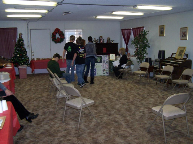 Ein Raum mit mehreren Stühlen, Tischen und Personen, einige stehen, einige sitzen. Auf den Tischen stehen Becher und Gefäße. Im Hintergrund eine Wand mit einem Strauß, Vorhang, Tür, Fotorahmen und Fenstern. Ein Weihnachtsbaum und ein Piano auf der rechten Seite, mit Röhrenleuchten an der Decke.