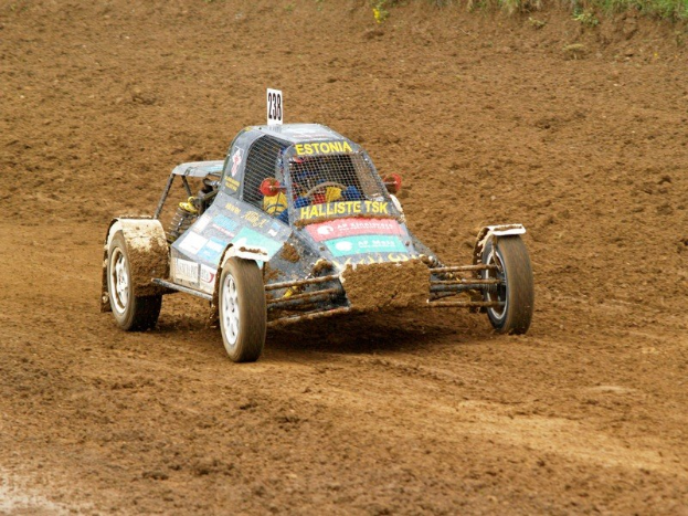 Ein Mann mit Helm fährt einen Buggy auf einem Schotterweg, umgeben von Pflanzen, mit sichtbarem Text auf dem Fahrzeug.