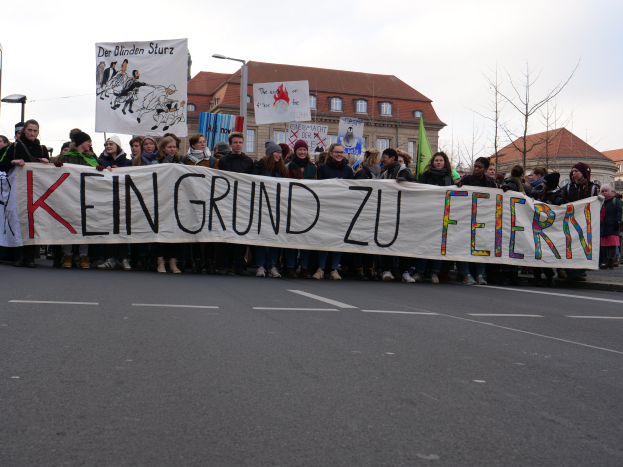 Eine Gruppe von Menschen, die auf der Straße stehen und ein Transparent mit der Aufschrift "Kein Grund zu Feiern" halten, vor dem Hintergrund von Straßenlaternen, Bäumen, Gebäuden und einem klaren blauen Himmel.