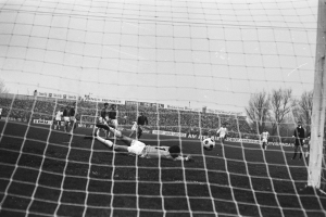 Ein Schwarz-Weiß-Foto eines Fußballspielers, der vor einem Tor einen Ball hält, umgeben von einer Gruppe von Menschen, einem Ball, einem Netz, einem Pfosten, einem Zaun, einer Fahne mit Text, Bäumen und einem bewölkten Himmel.