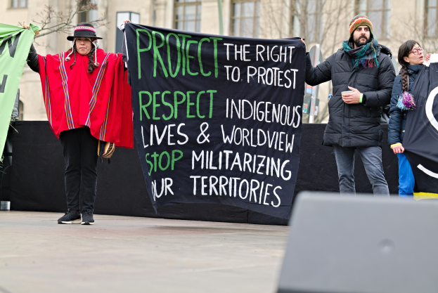 Gruppe von Menschen mit einem Banner mit der Aufschrift "Schutz des Rechts auf Demonstration Respektiere indigene Leben und Weltanschauung Stoppe die Militarisierung unserer Territorien" vor einem Gebäude mit Bäumen und einem bewölkten Himmel.