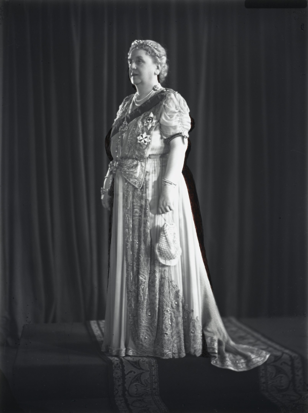 Schwarz-weißes Foto von Queen Elizabeth II auf einer Bühne mit einem Vorhanghintergrund, die eine königliche Robe mit feinen Details und einem langen Rock trägt, das Haar in einem eleganten Dutt frisiert, mit einem sanften Gesichtsausdruck zur Seite schauend.