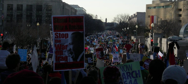 Menschen protestieren auf einer Stra√üe und halten Plakate in H√∂nden, mit Geb√§uden, B√§umen, Laternenm√§sten und dem Himmel im Hintergrund.