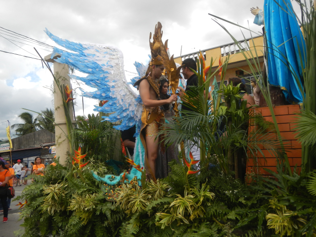 Eine Gruppe von Menschen geht eine Straße entlang neben einem Karnevalswagen, der mit Pflanzen und Blumen geschmückt ist, mit kostümierten Personen auf dem Wagen, Gebäuden, Bäumen und Strommasten im Hintergrund unter einem bewölkten Himmel.