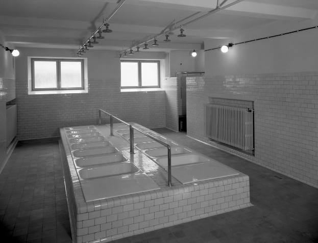 Schwarzes und weißes Foto eines öffentlichen Waschraums mit einer langen Reihe gefliester Waschbecken, Armaturen, Wände, Fenster, Lampen und Rohre bei natürlichem Licht.