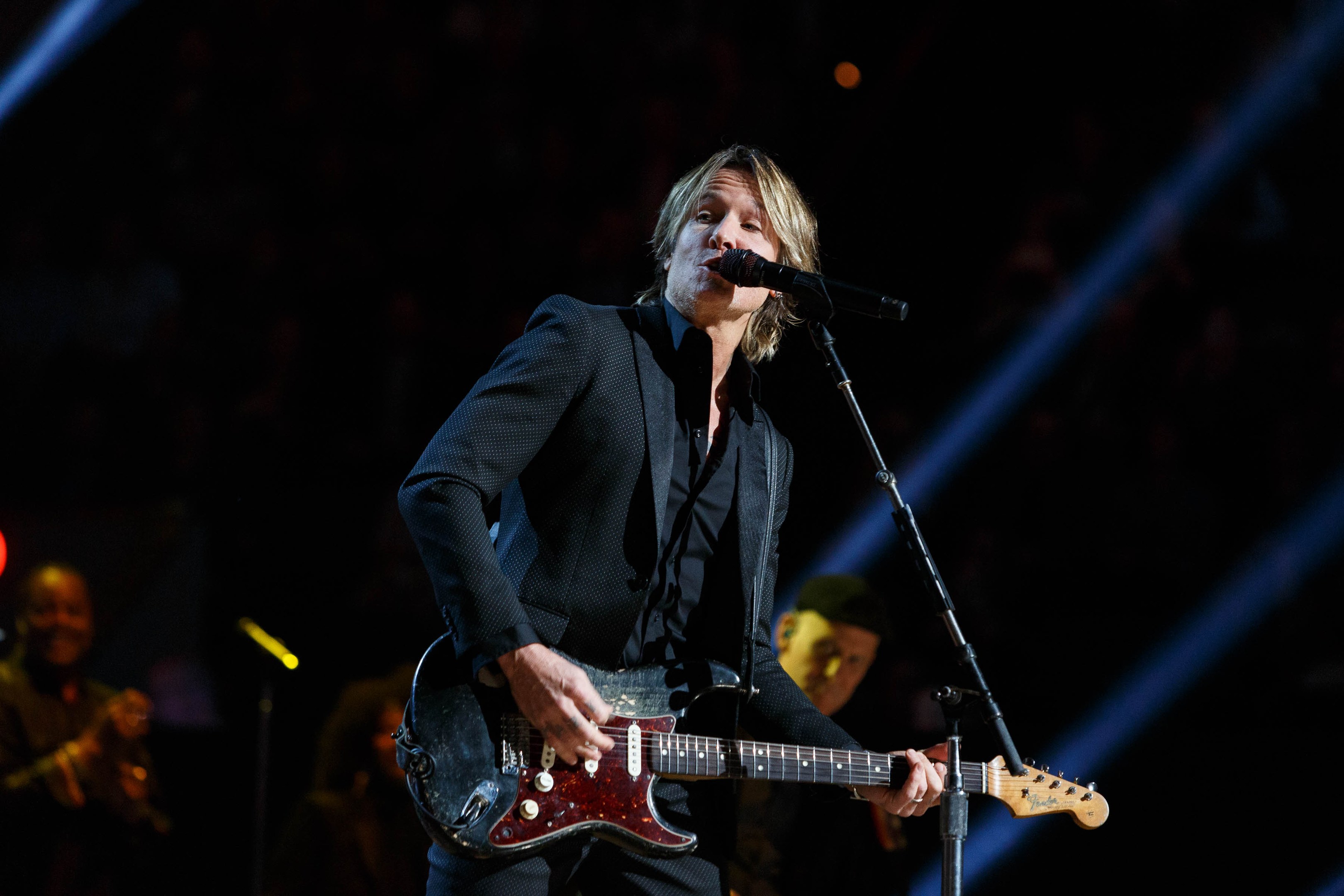 Keith Urban performing on stage at the Joint in Las Vegas, playing a guitar with a microphone in front of him and musicians in the background, under dark lighting.