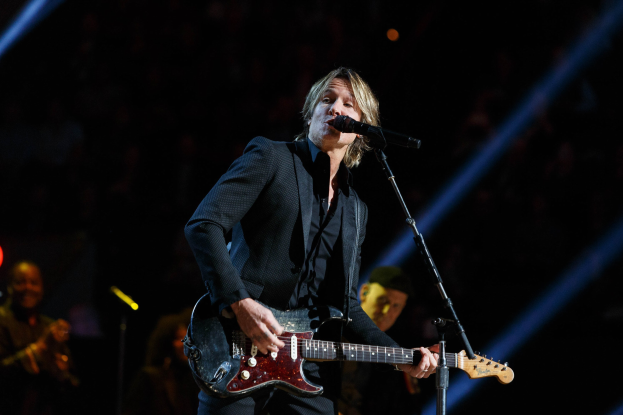Keith Urban performing on stage at the Joint in Las Vegas, playing a guitar with a microphone in front of him and musicians in the background, under dark lighting.