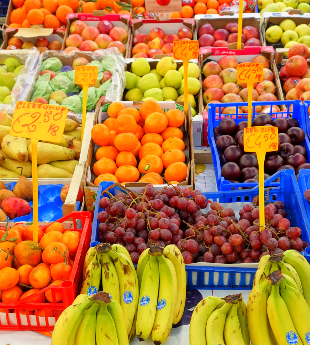 Ein Marktstand mit einer Auswahl an Obst und Gemüse, wie Bananen, Äpfeln, Orangen und Trauben, mit Preisschildern an jedem Artikel.