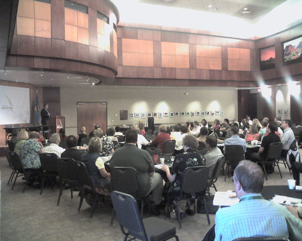 Ein Konferenzraum mit einem Bildschirm, einer Flagge daneben, einer Person hinter einem Rednerpult, einer Gruppe sitzender Menschen, verschiedenen Bildern an der Wand und einem Tisch mit einer Tasse und Papieren.