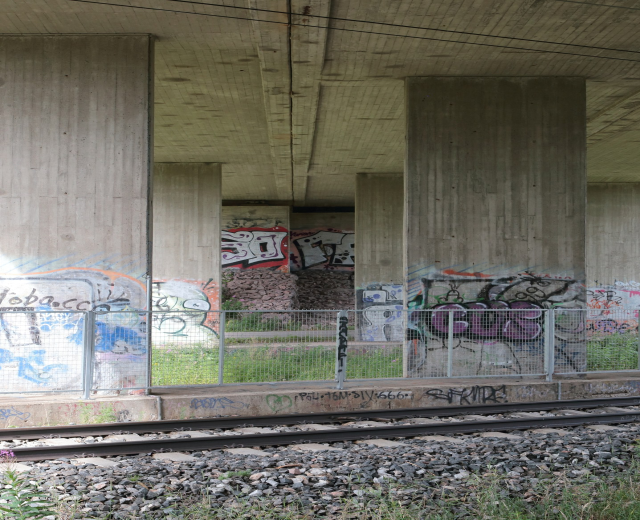 Schienenstrecke mit Graffiti auf einem Stein und einem grasbewachsenen Bereich, gesäumt von einem Zaun, Säulen, Oberleitungen und einem Dach.