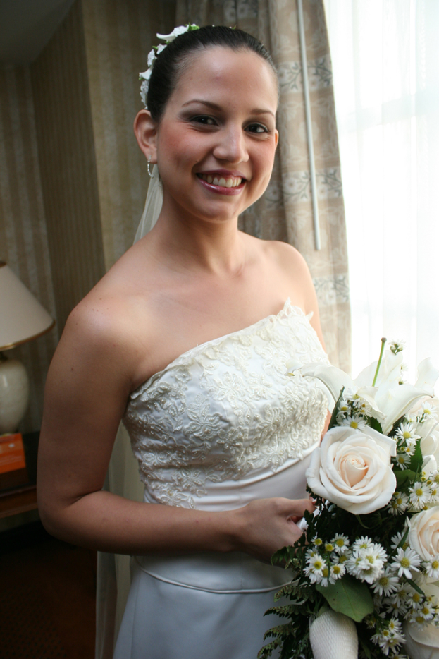 Eine Braut steht lächelnd da und hält einen Strauß, mit einem Vorhang und einer Wand im Hintergrund und einer Lampe auf einem Tisch links.