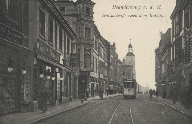 Ein altes Schwarz-Weiß-Foto einer Straßenlandschaft in Brandenburg, Deutschland, mit einer Tram auf Schienen, Menschen auf dem Gehweg, Gebäuden mit Fenstern, Schildern, Straßenlaternen und einem bewölkten Himmel.