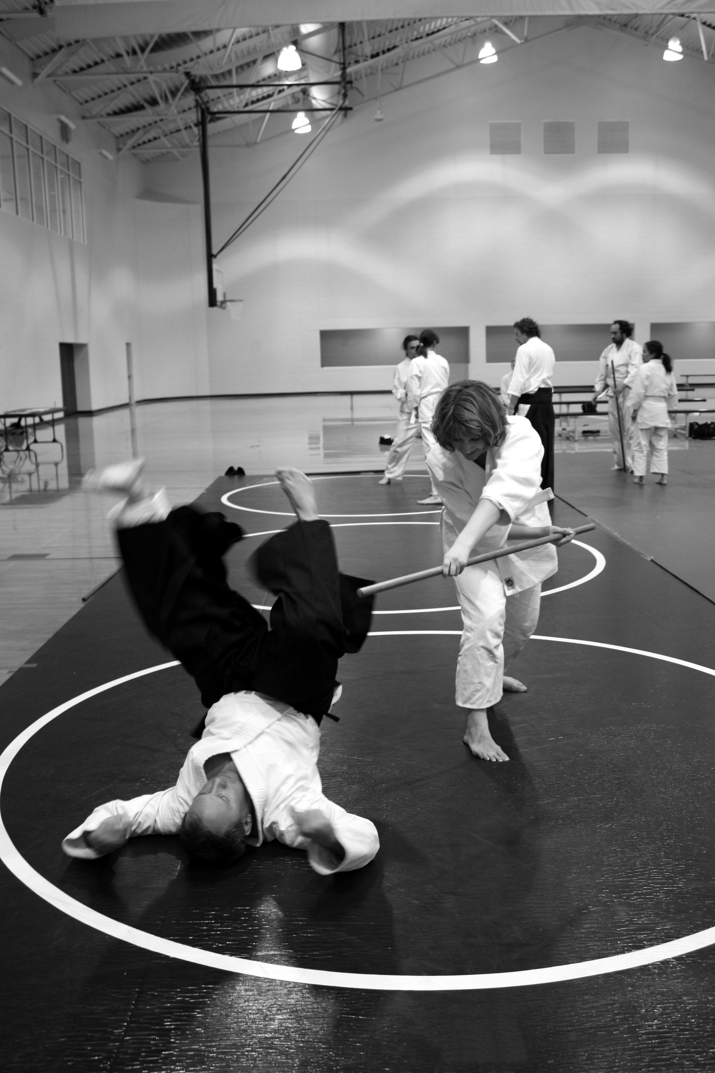 Zwei Personen üben Karate mit Stöcken in einer Turnhalle, während andere zuschauen, mit Leuchten entlang der Wand im Hintergrund.