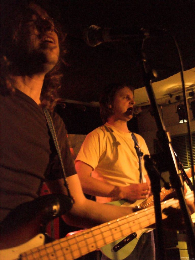 Eine Person in einem weißen Hemd singt in ein Mikrofon und spielt eine grüne Gitarre, während sie neben einer anderen Person in einem schwarzen Hemd, mit Brille und einer schwarzen Gitarre steht, mit Scheinwerfern und anderen Instrumenten im Hintergrund.