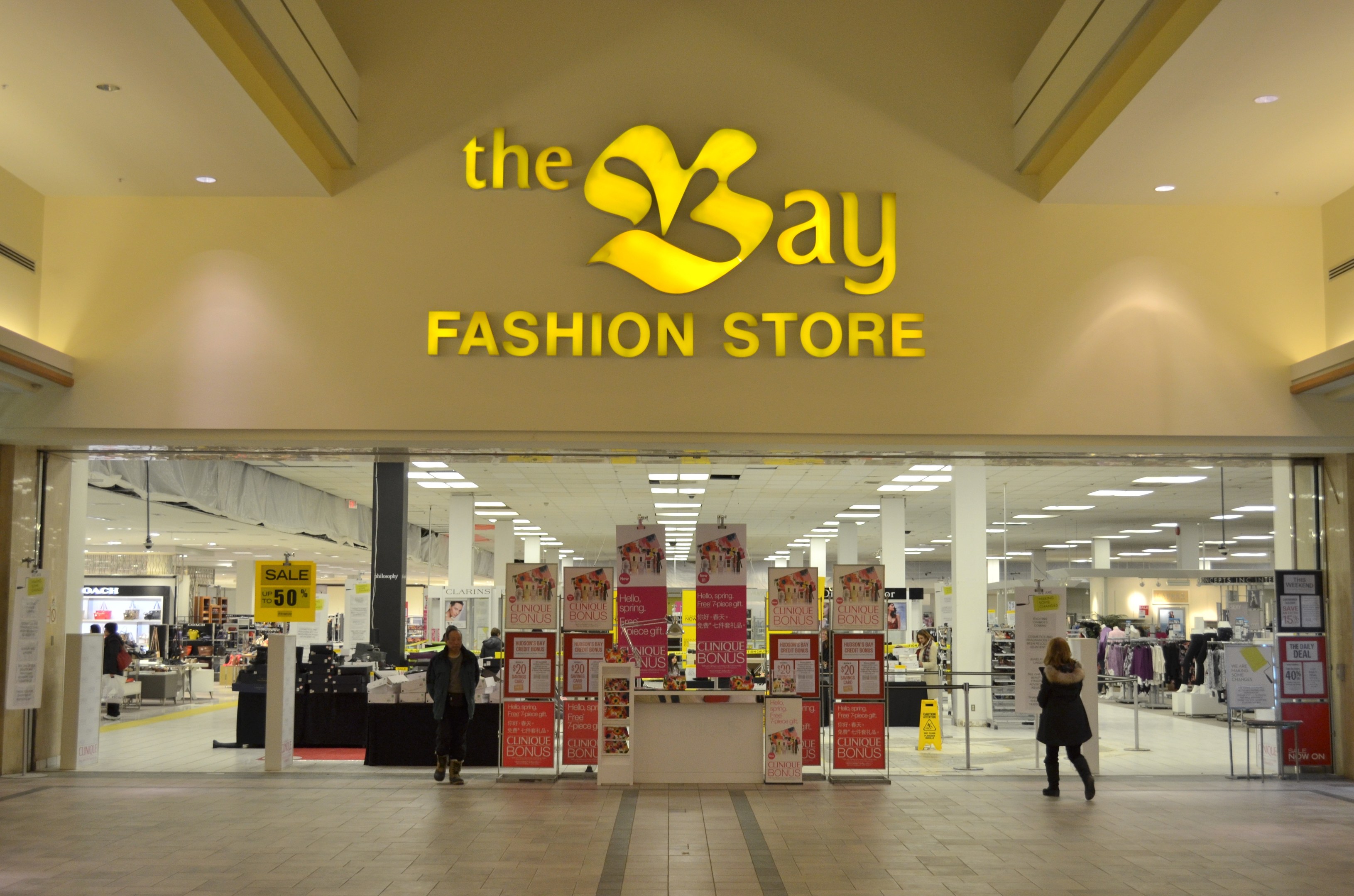 Außenansicht eines Bay Fashion Store in einem Einkaufszentrum mit Einkäufern, Wandtexttafeln, Säulen, Deckenleuchten und Hintergrundauslagen.