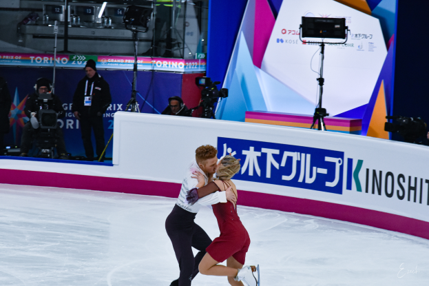 Ein Paar umarmt sich beim Eislaufen auf einer Eisfläche, während Menschen mit Kameras in der Nähe stehen und ein Schild und Lichter im Hintergrund zu sehen sind.