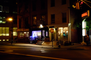 Eine Stadtansicht mit Geb├Ąuden, beleuchteten Gesch├Ąften, Treppen, B├Ąumen, einer Stra├če, einem Fahrrad und einer Ampel.