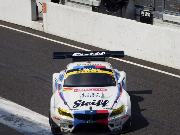 Ein BMW M6 GT3 Rennwagen auf einer Rennstrecke mit einer Wand, einem Maschengitter und anderen Objekten im Hintergrund.