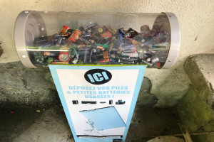 Ein Mülltonnen voller Batterien auf einem Gehweg, mit einem Plakat mit Text an der Wand dahinter und Treppen auf der rechten Seite.