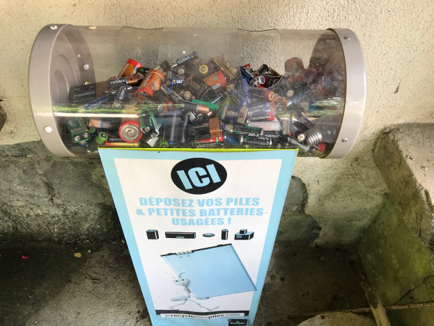 Ein Mülltonnen voller Batterien auf einem Gehweg, mit einem Plakat mit Text an der Wand dahinter und Treppen auf der rechten Seite.