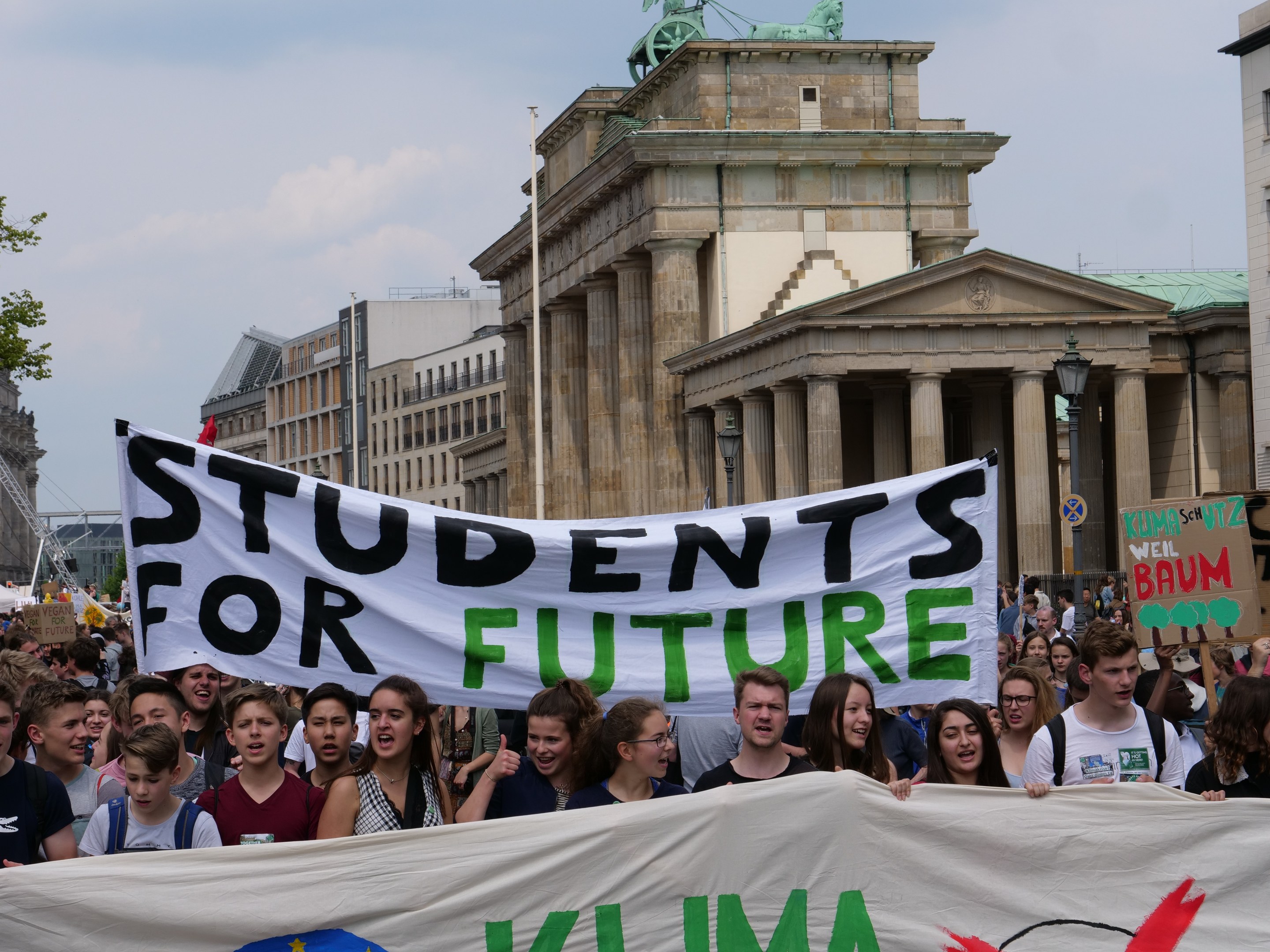 Schüler in Berlin marschieren mit buntem "Students for Future"-Schild gegen Gebäude, Bäume und Himmel.
