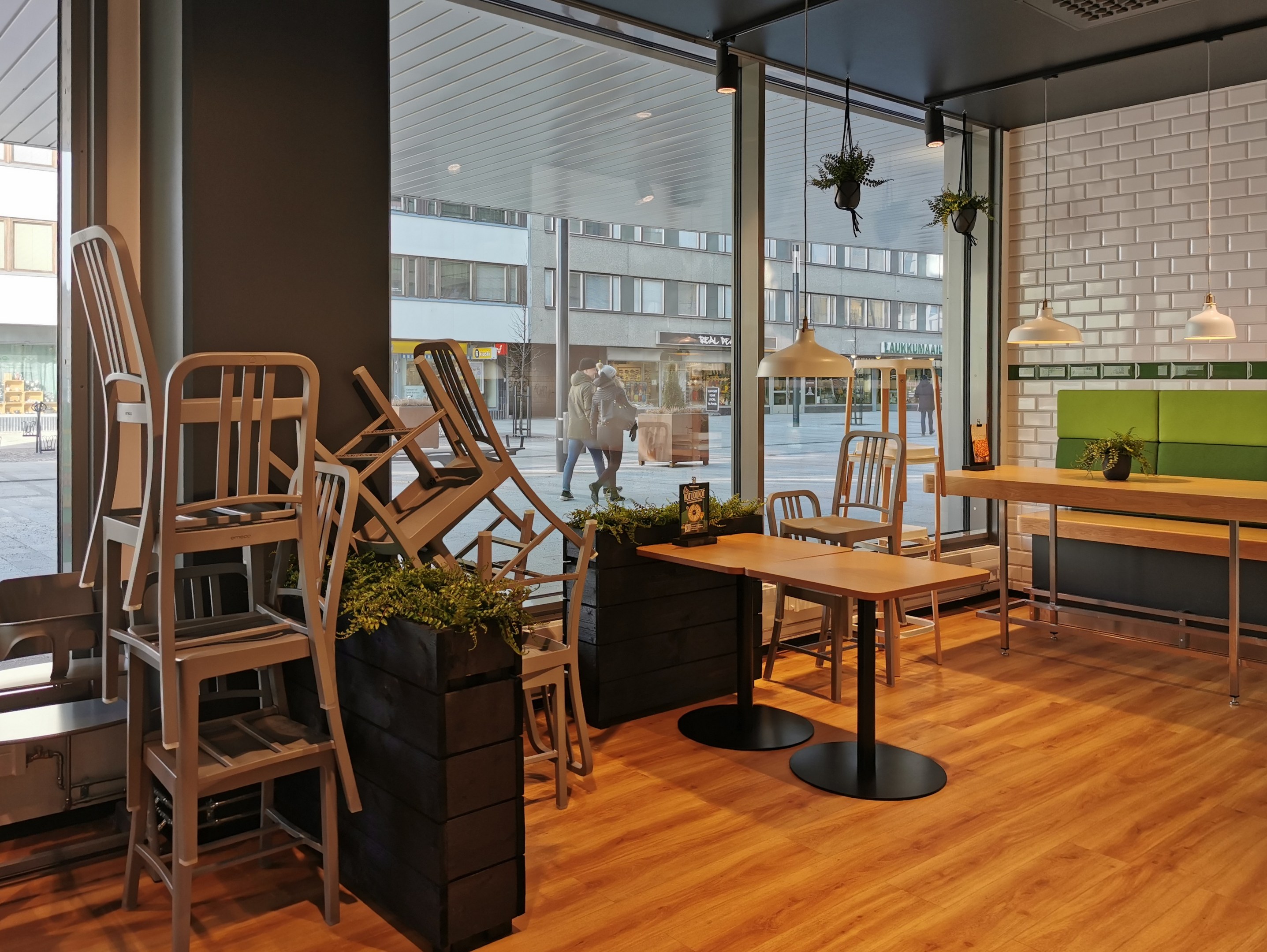 Außenansicht eines Berliner Restaurants mit Holztischen und -stühlen, großem Fenster, Pflanzen, Beleuchtung und Blick auf eine Straße mit Fußgängern, Gebäuden und städtischen Elementen.