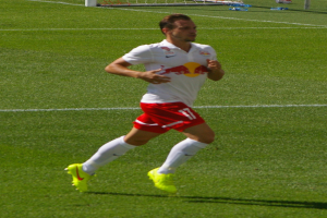 Ein Mann in einem weißen Hemd und roten Shorts läuft auf einem Fußballfeld, im Hintergrund ist ein Tor zu sehen und ein paar Menschen stehen in der Nähe, die Fußballschuhe tragen.