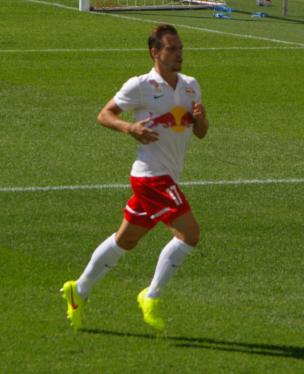 Ein Mann in einem weißen Hemd und roten Shorts läuft auf einem Fußballfeld, im Hintergrund ist ein Tor zu sehen und ein paar Menschen stehen in der Nähe, die Fußballschuhe tragen.