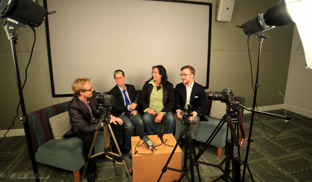 Eine Gruppe von drei Personen sitzt auf einem Sofa und gibt ein Interview, wobei eine Person links in einem schwarzen Mantel und Hose sitzt, eine Person in der Mitte einen Mikrofon haltenden Mantel und eine Krawatte trägt und eine Person rechts lächelt. Hinter ihnen befindet sich ein Projektionsschirm, ein Mikrofonrekorder auf dem Tisch in der Mitte und Kameras mit großen Stativen auf beiden Seiten.