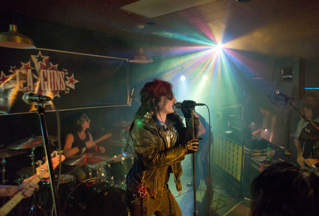 Eine Frau in einer schwarzen Jacke singt auf der Bühne in ein Mikrofon, während ein Mann mit Sonnenbrille und Stirnbinde hinter ihr ein Instrument spielt; ein 'LA Guns'-Plakat und Diskolichter sind im Hintergrund zu sehen.