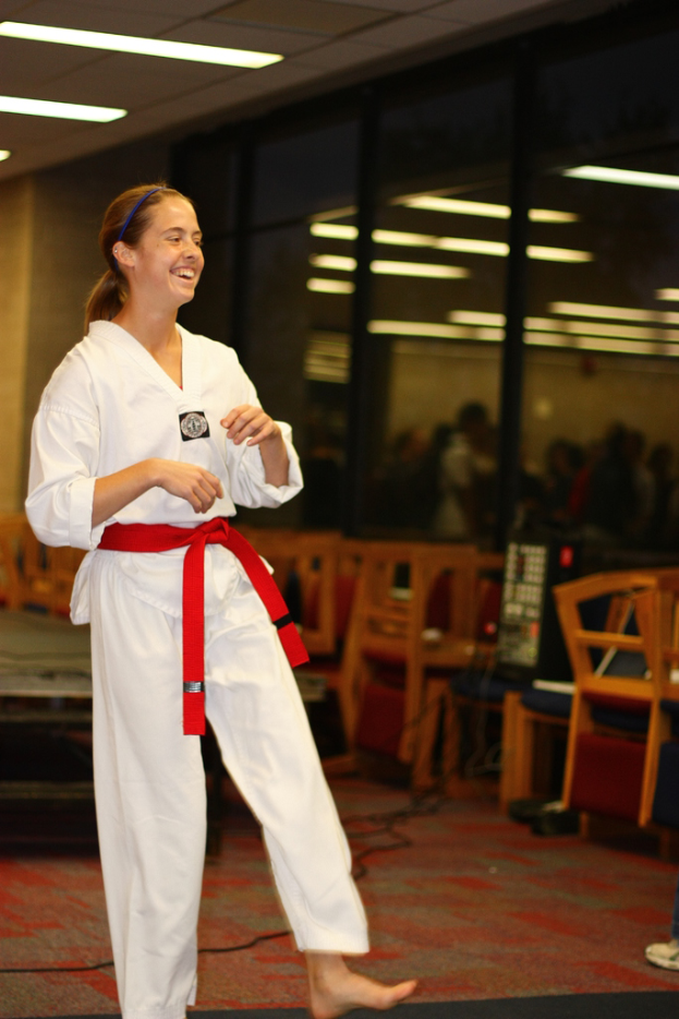 Ein Mädchen in einem Karate-Outfit mit einem roten Gürtel lacht, schaut auf etwas, mit Stühlen, einer Musikanlage und ein paar Menschen hinter ihr, an einer Wand mit Lichtern.