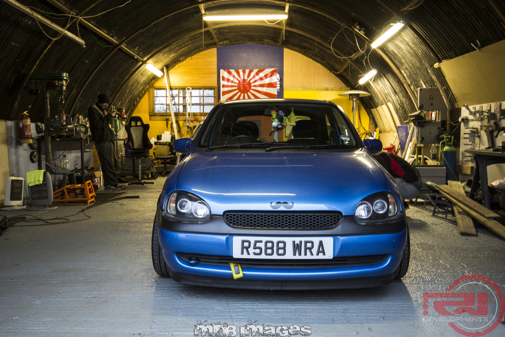 Ein blaues Auto steht in einem Schuppen mit einer Flagge an der Rückwand, Deckenlampen und Maschinen, Werkzeugen und Zubehör auf beiden Seiten; ein Mann steht links vor der Wand.