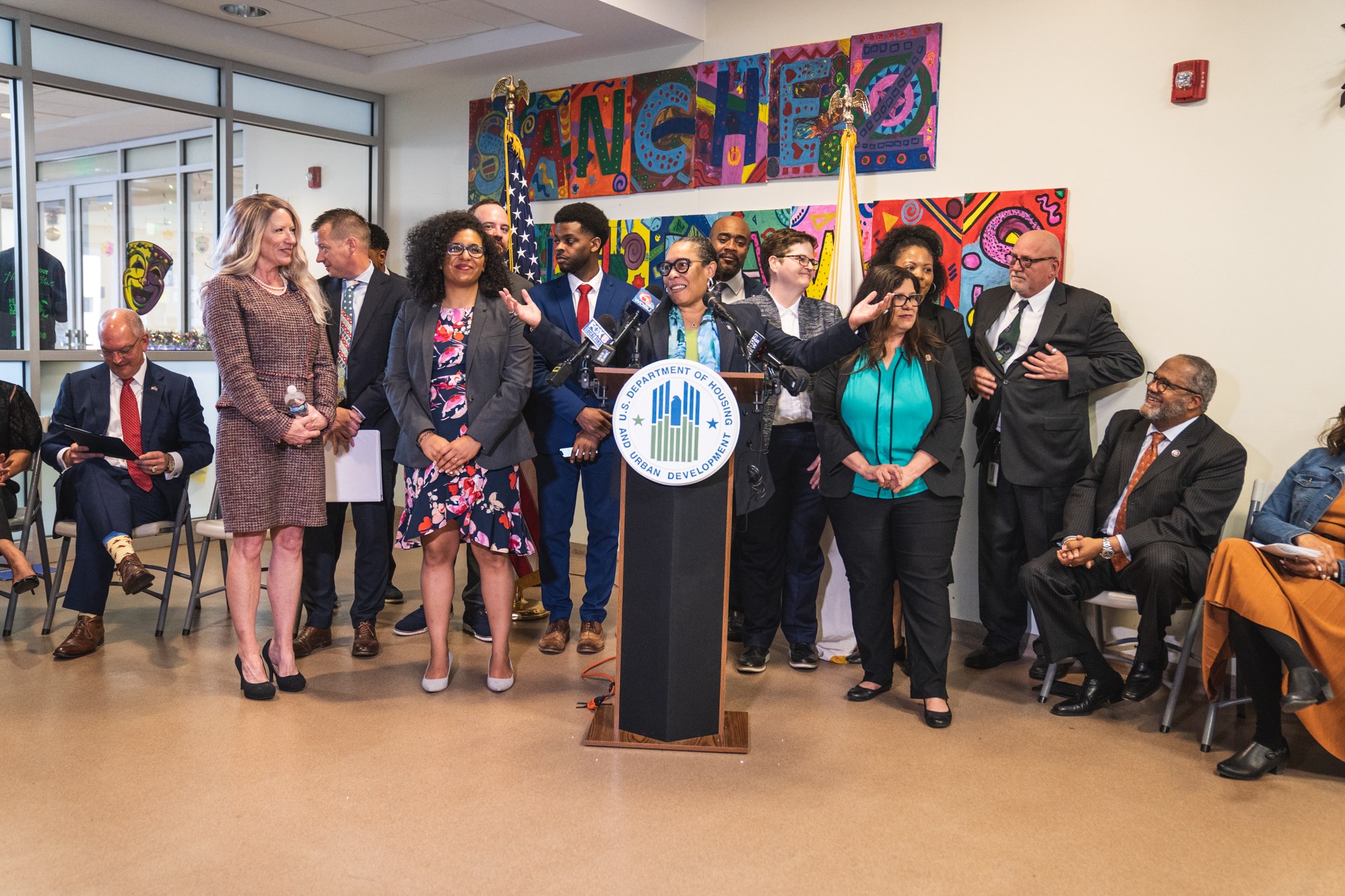 Eine Gruppe von Menschen steht vor einem Podium mit einem Mikrofon und einer Tafel mit dem Text "Stadtrat von Sancho Stadt joins City Council". Andere sitzen auf Stühlen oder stehen in der Nähe; Flaggen, ein Feuerlöscher und Deckenleuchten sind an der Rückwand zu sehen.