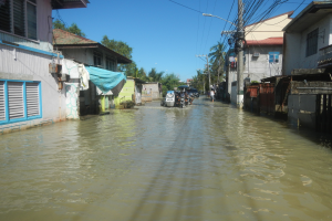 Eine überflutete Straße in einem kleinen Ort mit Menschen, die durch das Wasser gehen, Gebäude mit Fenstern und Türen auf beiden Seiten, Strommasten mit Drähten, Bäume und einen klaren Himmel im Hintergrund.