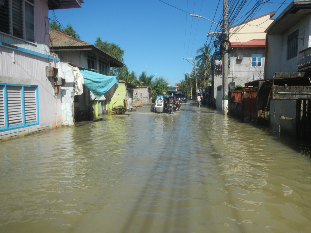 Eine überflutete Straße in einem kleinen Ort mit Menschen, die durch das Wasser gehen, Gebäude mit Fenstern und Türen auf beiden Seiten, Strommasten mit Drähten, Bäume und einen klaren Himmel im Hintergrund.
