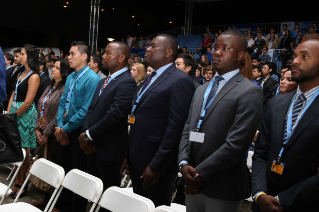 Männer in Blazern und ID-Karten stehen auf einem Boden mit Stühlen vor dunklem Hintergrund mit Absperrgittern und Eisenpfeilern.